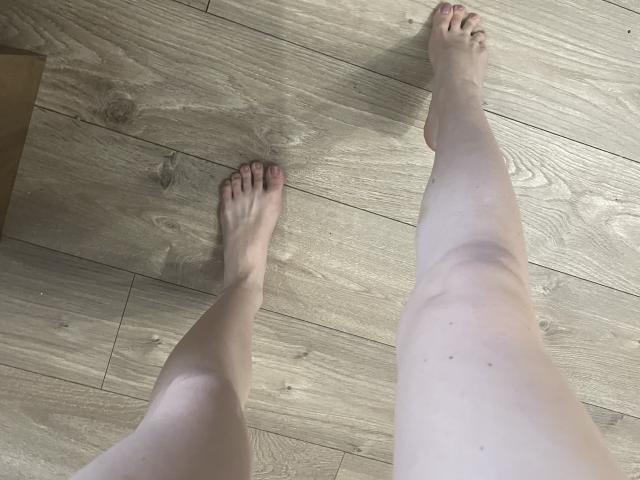 Les jambes et les pieds pâles de RozalinaPleasure sont posés sur un plancher en bois, ses orteils pointant légèrement vers l'intérieur. Sa peau rosée présente de petites taches brunes sur ses cuisses..