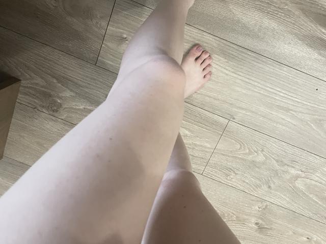 RozalinaPleasure's bleke benen waren gekruist bij de enkels, waarbij haar tenen zichtbaar waren op een houten vloer. Haar blote dijen en gladde huid bevinden zich op de voorgrond..