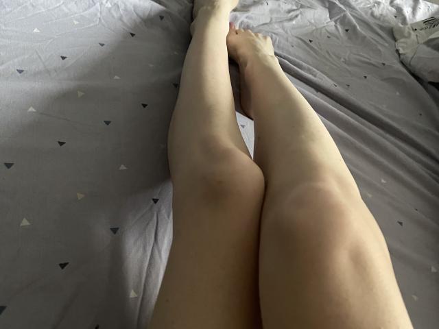 As pernas pálidas e esguias de RozalinaPleasure estão cruzadas em uma cama cinza com pequenos padrões triangulares. Seus dedos estão apontados e a cama tem leves rugas..