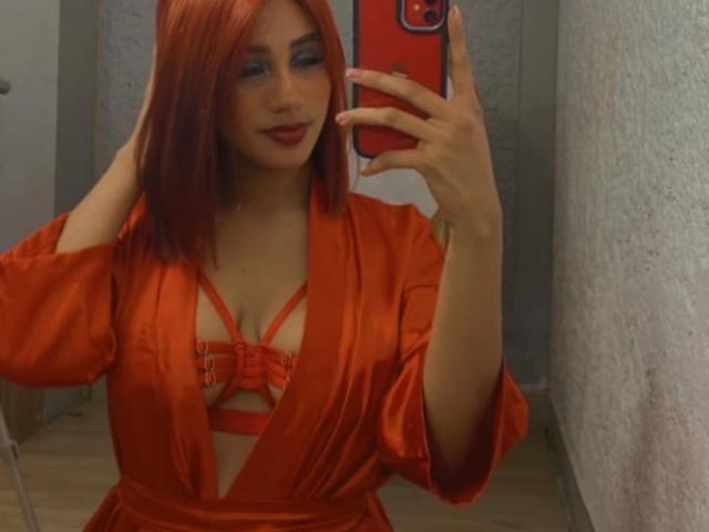ChloeAnnistone, avec des cheveux roux, portant une robe en soie orange et de la lingerie rouge à bretelles fines, prend un selfie devant un miroir avec son téléphone, montrant ses seins..