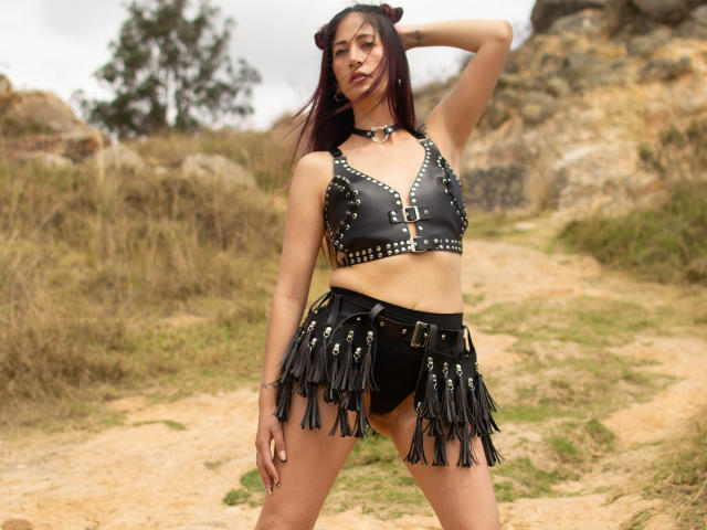 CateVanss hat dunkelbraunes Haar in zwei Duttfrisuren, trägt ein schwarzes Leder-Crop-Top mit Nieten und einen Rock mit Fransen, schwarze Unterwäsche und eine Halskette. Sie steht draußen..