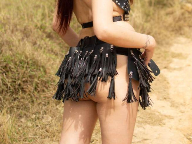 Het blote achterwerk van CateVanss is bedekt met een zwarte, gefranjde rok met leren bandjes. Ze staat op een onverhard pad met gras, en haar tatoeage is zichtbaar op haar rechterarm..