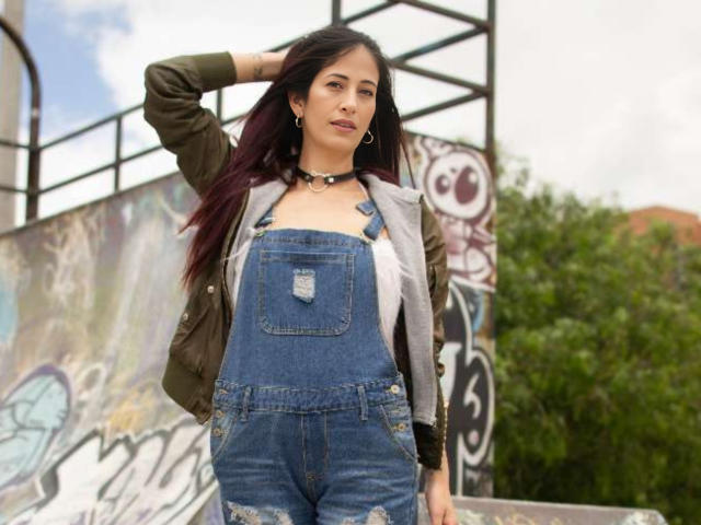 CateVanss tiene el pelo oscuro, lleva una chaqueta verde, una sudadera gris y unos pantalones vaqueros azules con un bolsillo rasgado, está de pie con una mano en el pelo y mirando a la cámara..