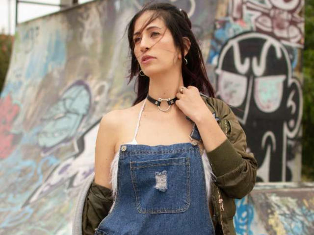 CateVanss with dark hair, wearing a green jacket, denim overalls with a pocket tear, and a black choker, holding the collar, standing in front of graffiti.