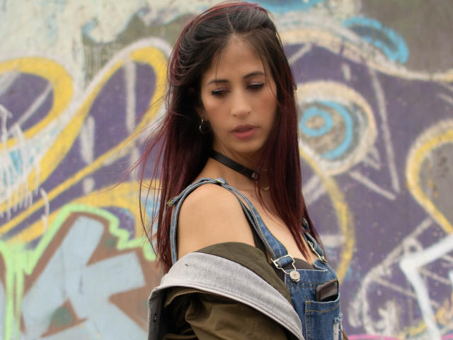 CateVanss con lunghi capelli rosso scuro, indossa una tuta in denim, una camicia verde, una collana nera, guarda in basso, con una spalla scoperta, espressione facciale delicata, un muro con graffiti sullo sfondo..