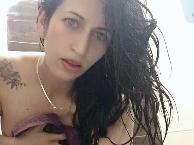 CateVanss, con el cabello negro y húmedo, sosteniendo una toalla morada cubriendo sus senos, tiene un tatuaje floral en el hombro derecho y un collar de plata, mirando a la cámara..