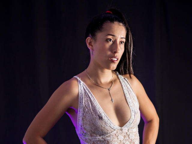 KatherinParmer hat dunkles Haar zu einem Pferdeschwanz gebunden und trägt ein weißes Spitzenkleid mit tiefem Ausschnitt, das ihre Brüste zeigt. Sie trägt eine silberne Halskette und steht vor einem dunklen Hintergrund..