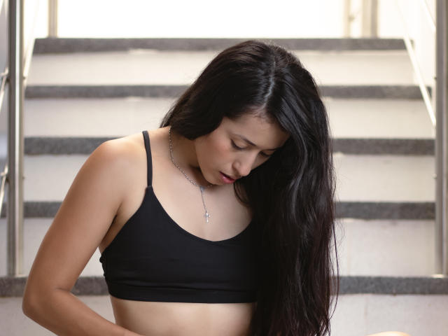 KatherinParmer has long black hair, a black sports bra, and a silver necklace, looking down with a sad expression, standing in front of gray stairs.