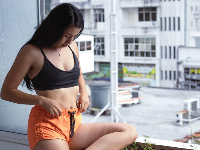 KatherinParmer, avec de longs cheveux noirs, un petit soutien-gorge de sport noir et un short orange avec de la dentelle noire, assise près d'une fenêtre, regardant vers le bas, les doigts près de sa taille..