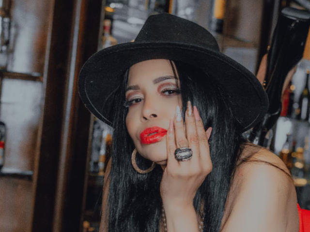AngelinaDeleon with black hat, red lipstick, and long black hair, touching her face with a ringed hand, wearing hoop earrings, in a bar with blurred background.