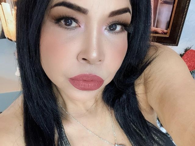 AngelinaDeleon has black hair, brown eyes, and pinkish skin. She's wearing red lipstick and a silver necklace, with a black top. Her tits are slightly visible.