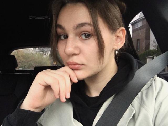 SofiaNash has a cute face with light brown eyes, small nose, and slightly pouty lips. She's wearing a gray jacket, black scarf, and seatbelt, with brown hair in a bun.