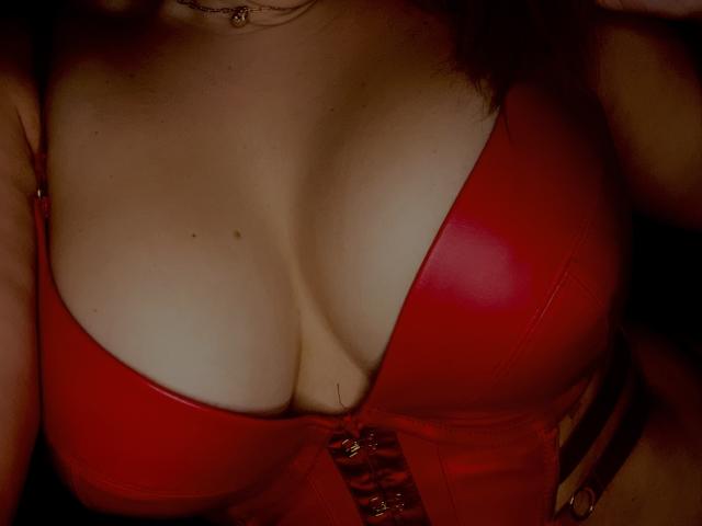 LucyHottX's tits in a shiny red leather bra, cleavage deep, small mole on her left, bra clasps in front, dark shadowy background, subtle necklace visible.