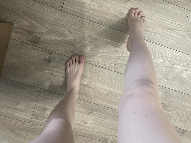 As pernas e os pés pálidos de RozalinaPleasure estão em um chão de madeira, com os dedos ligeiramente apontados para fora. Suas unhas dos pés estão pintadas de um vermelho escuro..