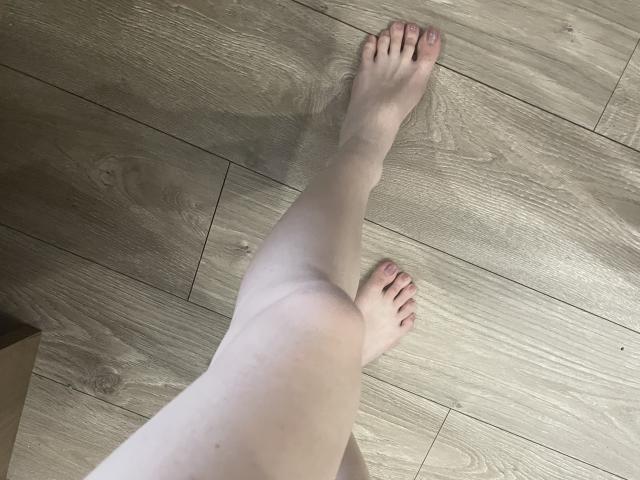 Le gambe e i piedi pallidi di RozalinaPleasure sono incrociati su un pavimento di legno, mostrando le dita dei piedi e le piante leggermente sporche. La sua pelle è liscia, con qualche piccola imperfezione..