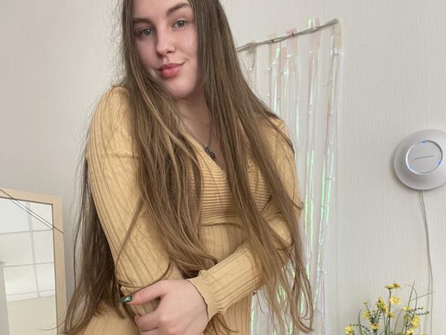 Dooris with long brown hair, cute face, and pouty lips, wearing a ribbed, beige, long-sleeve top, arms crossed, standing, showing a slight smile, in a casual setting.