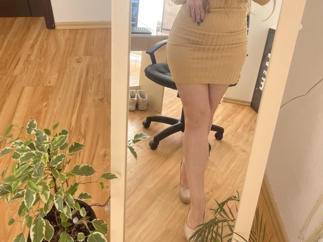 Doris in a tight, ribbed, beige mini-skirt and beige heels, standing with her ass and legs visible, reflecting in a mirror, next to a potted plant and office chair.