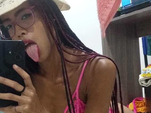 CanelaHott69, avec des tresses roses, des lunettes roses, la langue tirée et un chapeau, prend un selfie devant un miroir avec son téléphone, montrant de petites seins et un corps mince, à la peau foncée..