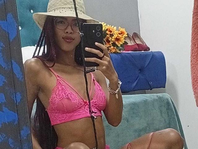 CanelaHott69, com pele escura, tranças longas, usando óculos, um sutiã de renda rosa e um chapéu de palha, segurando um telefone, mostrando seus pequenos seios e nádegas planas, em uma selfie no espelho..