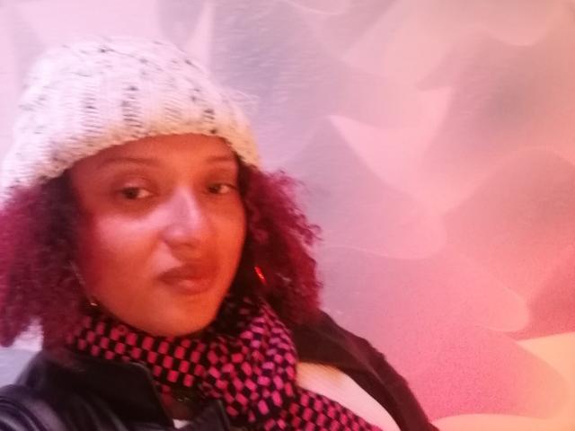 VioletaSwitch with curly purple hair, white knit hat, black leather jacket, and pink checkered scarf, showing a subtle pout. Boob cleavage visible. Soft, pinkish light.