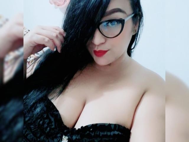 Briana69x, avec des cheveux noirs, des lunettes, du rouge à lèvres rouge, un haut en dentelle noire sans bretelles, montrant de grandes seins, la main droite touchant ses cheveux, fond blanc, léger sourire, mignonne, pulpeuse, sexy..
