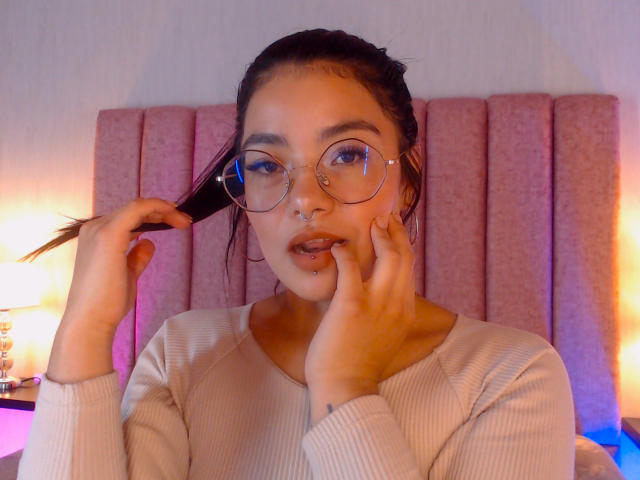 Emma Ruiz, usando óculos, segurando o cabelo, tem um piercing no nariz e nos lábios, vestindo uma blusa bege texturizada, tocando o rosto, em um quarto com iluminação suave..