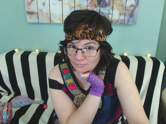 EverlyRays with glasses, leopard headband, purple glitter glove, thinking with chin on hand, striped black and white couch, colorful beaded necklace, short black hair, light skin, smiling.
