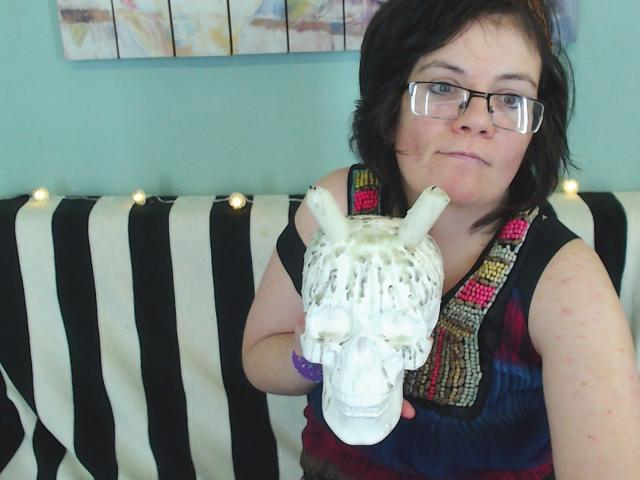 EverlyRays with glasses, short black hair, and a purple bracelet, holding a white, painted skull with horns, wearing a colorful sleeveless top, sitting on a black-and-white striped couch.