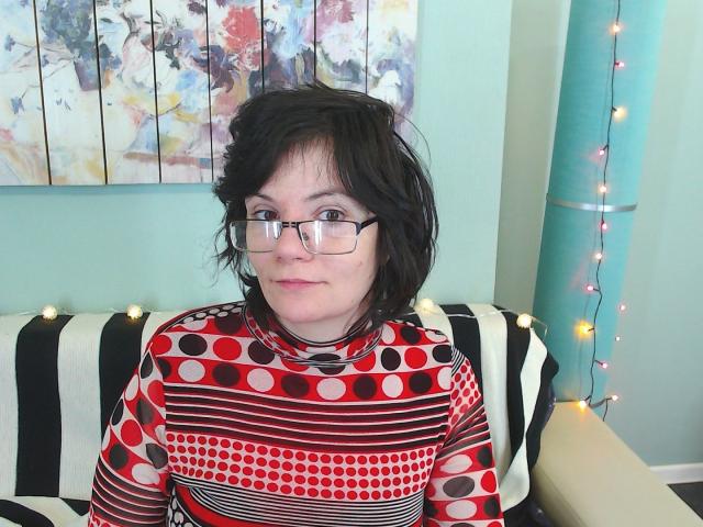 EverlyRays with short black hair, glasses, and a red polka dot and striped turtleneck, sitting on a black and white striped couch, looking slightly confused.