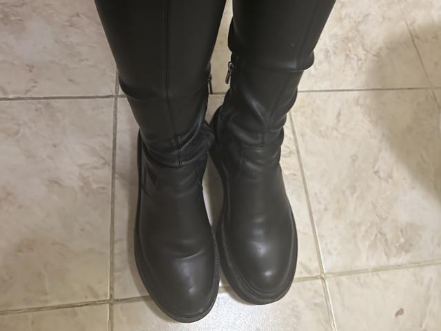 As botas de couro preto da DiLaria estão em pé sobre um piso de azulejos bege. As botas estão ligeiramente desgastadas, com vincos visíveis e um zíper na lateral..