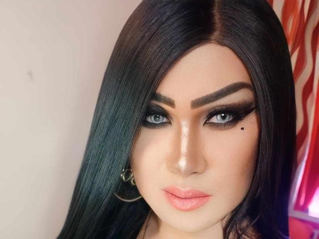 BornStarTS has dark, straight hair, blue eyes, thick eyebrows, long eyelashes, and a small black mole on her right cheek. She's wearing pink lipstick and gold hoop earrings.