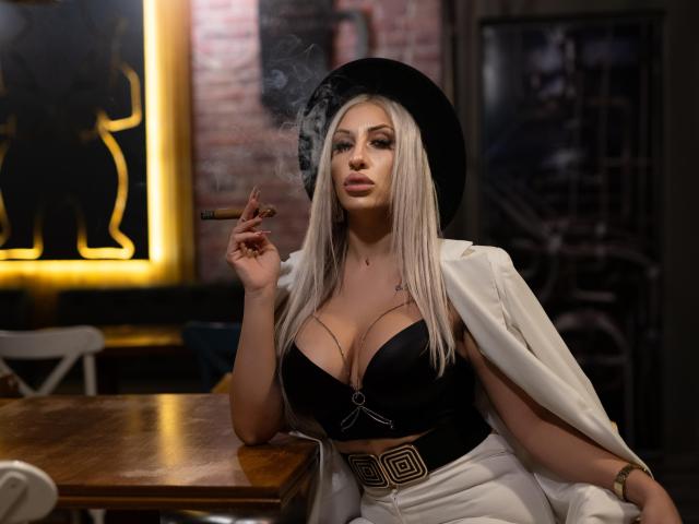 VickiSpices with long blonde hair, black hat, white jacket, black bra showing big tits, smoking, holding cigarette, white pants, sitting at wooden table, looking at camera.