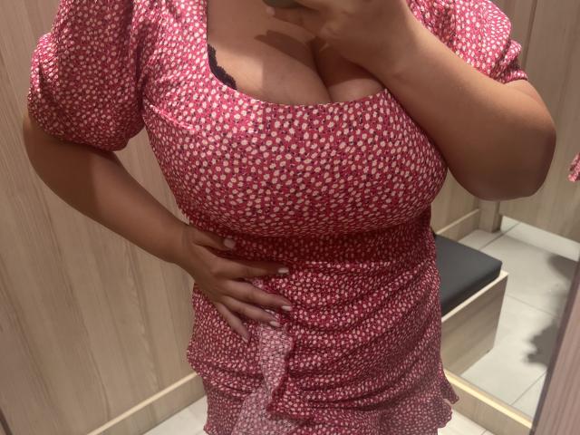 Barbarica indossa un vestito rosso con pois bianchi, mostra il suo seno prosperoso nello specchio, con una mano appoggiata al fianco, reggiseno nero visibile, pareti di legno marrone sullo sfondo..