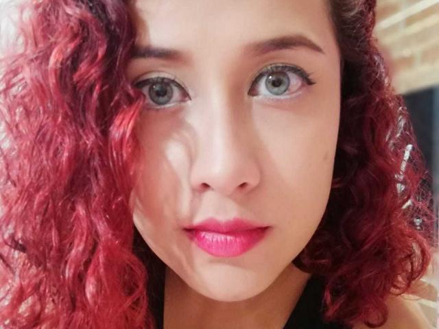 AbbyElecktra ha i capelli rossi ricci, gli occhi grigi e il rossetto rosa. Il suo viso è al centro dell'immagine, mostrando il naso, le sopracciglia e un leggero sorriso. Indossa abiti neri..