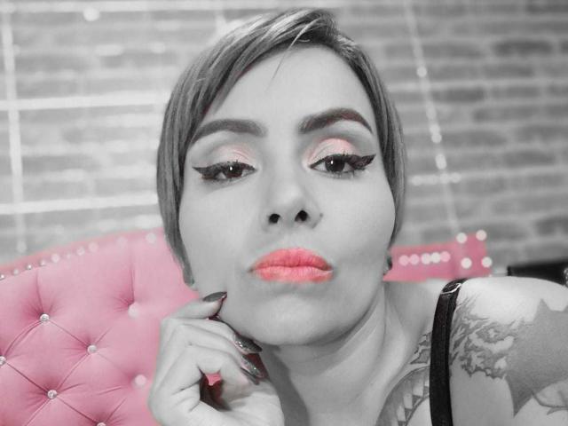 AbbyElecktra con cabello corto, sombra de ojos rosa y lápiz labial rojo, haciendo pucheros con la mano cerca de la cara, tatuaje negro en el hombro, silla rosa con borlas, fondo en escala de grises..