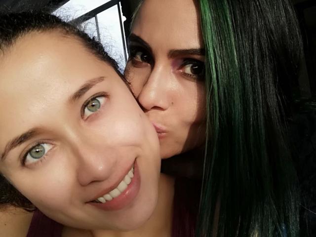 AbbyElecktra mit schwarz-grün meliertem Haar küsst AbbyElektras Wange und zeigt dabei ihre vollen Lippen und das violette Augen-Make-up. AbbyElecktra lächelt, hat blaue Augen, trägt kein Make-up und hat lockiges, braunes Haar..