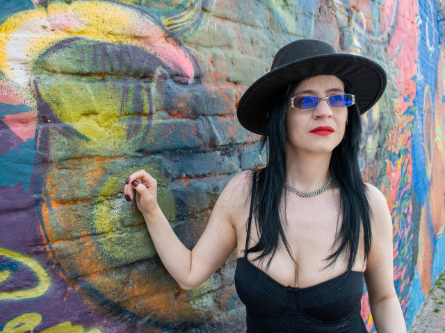AlanaMoneth with black hat, blue glasses, red lipstick, black strapless dress, and necklace, standing against colorful graffiti wall, showing her tits, black nails, and confident look.