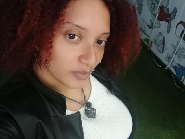 VioletaSwitch with red curly hair, light brown skin, and small nose ring, wearing a black leather jacket over a white top, necklace with a black pendant, looking at camera.