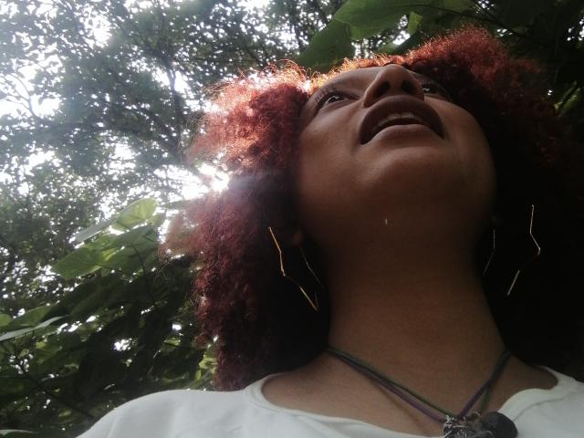 VioletaSwitch has dark brown skin, curly red hair, and is looking up with her mouth slightly open. She's wearing a white shirt and a necklace. Sunlight filters through leaves.