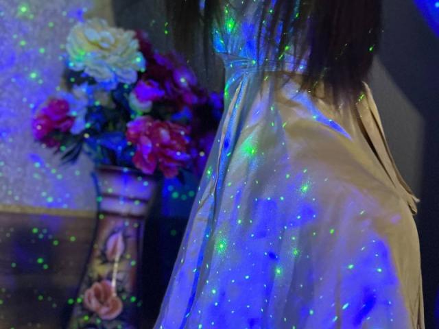 CheriLady portant une robe brillante, bleu-vert et pailletée, le dos à la caméra, avec un vase de fleurs en arrière-plan flou. Ses cheveux bruns sont partiellement visibles..