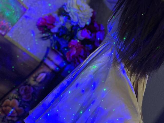 CheriLady steht mit dem Rücken zur Kamera, sie trägt ein glitzerndes blaues und weißes Kleid mit leuchtenden grünen Partikeln, und ihr braunes Haar ist leicht sichtbar. Im Hintergrund sind Blumen zu sehen..