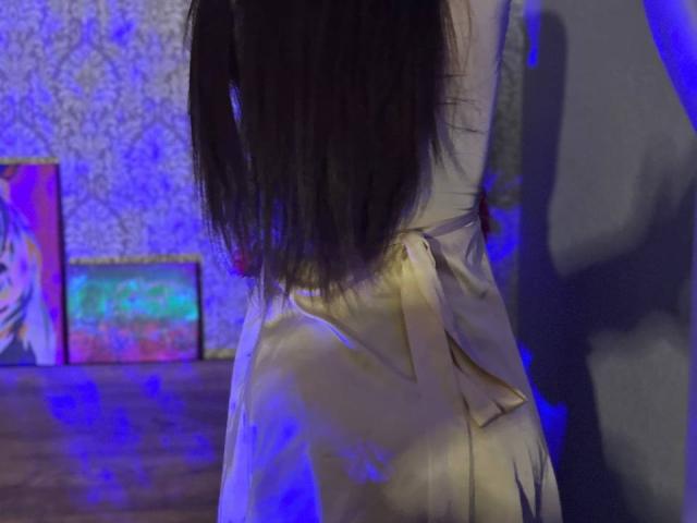 CheriLady est de retour, avec ses longs cheveux noirs, vêtue d'une robe beige en soie avec un nœud, debout dans une pièce éclairée en bleu, avec une ombre sur le mur, et des peintures colorées en arrière-plan..