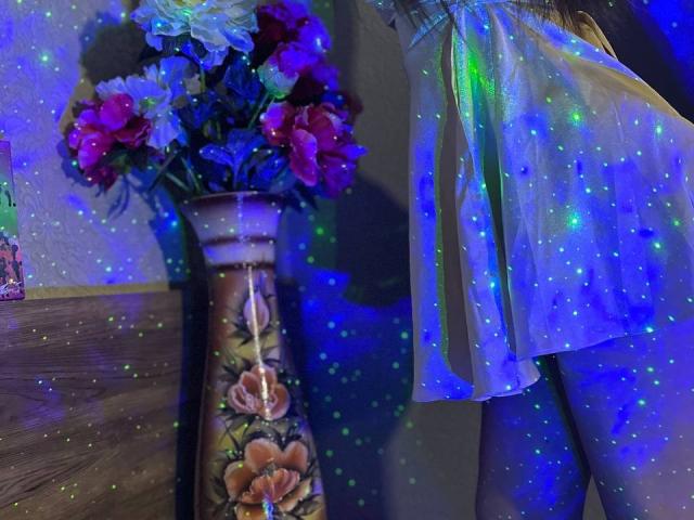 De glanzende jurk van CheriLady, met blauw-groene glitters, die haar achterwerk laat zien, staat naast een vaas met roze en witte bloemen, die oplichten onder blauw licht..