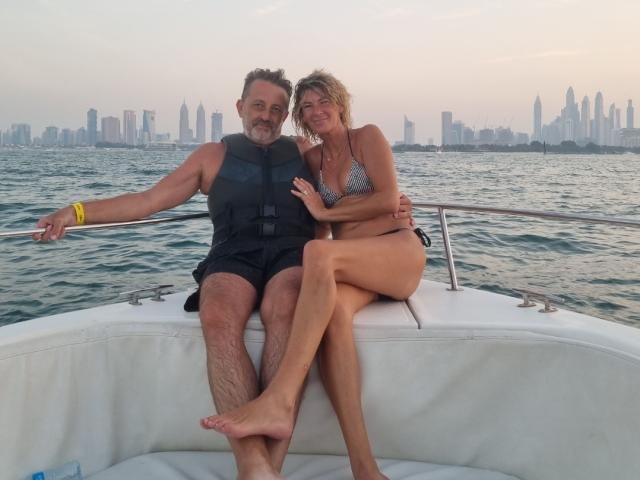 Um casal Angeel sentado em um barco, AngeelCouple (com seios em um biquíni cinza) com pele bronzeada, cabelo loiro, e AngeelCouple (com barba, colete preto, pulseira amarela) com pele clara, sorrindo..