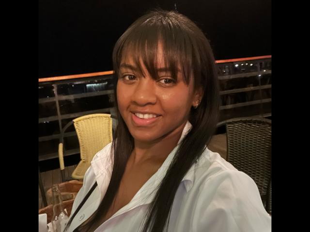 TiffanySammy has dark brown skin, long black hair with bangs, and is smiling at the camera. She's wearing a white shirt, with a glass and chair in the background.