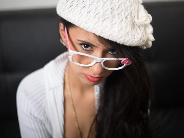 XiaQueen met donker haar, een witte gebreide muts, roze en witte bril, rode lippenstift, een witte geribbelde top, een gouden ketting en oorbellen, die omhoog kijkt met een lichte glimlach..