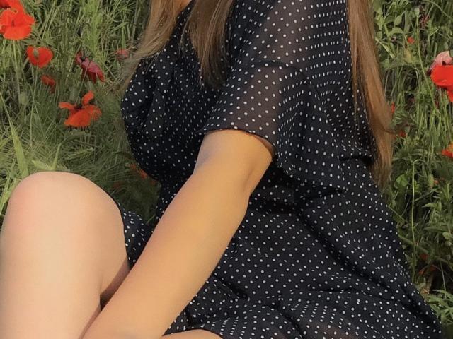 NutaAlisson in a black polka dot dress, sitting with knees up, surrounded by red poppies and green foliage. Long brown hair, dress with white dots, sunlight.