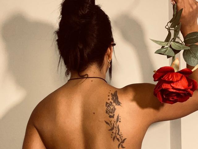 Amberv's back, dark hair in a messy bun, holding a red rose, tattoo of roses and leaves on her upper back, tan skin, shadow on white wall.