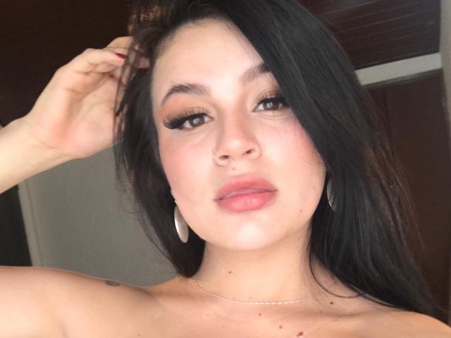 ShantallParis with dark, straight hair, brown eyes, and pink lips, wearing hoop earrings, a necklace, and red nail polish, touching her hair with a relaxed expression.