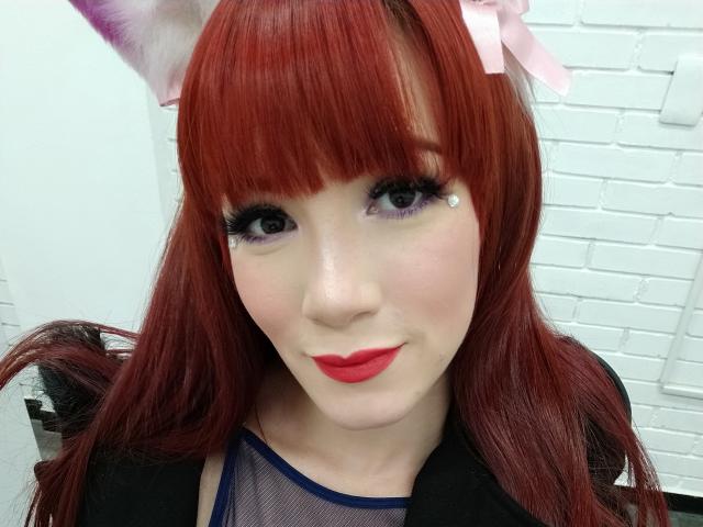 Akemi Haishi mit roten Haaren, weißen Katzenohren, lila Lidschatten, schwarzem Eyeliner, rotem Lippenstift und einem pinken Schleifchen, die ihr süßes Gesicht zeigt und lächelt. Sie trägt ein durchsichtiges blaues Oberteil..
